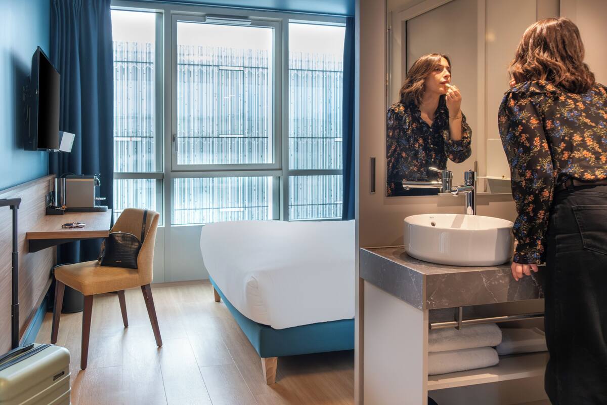 Una habitación de hotel moderna con cama, escritorio, silla, ventana grande, baño con lavamanos y espejo, y televisor en la pared.
