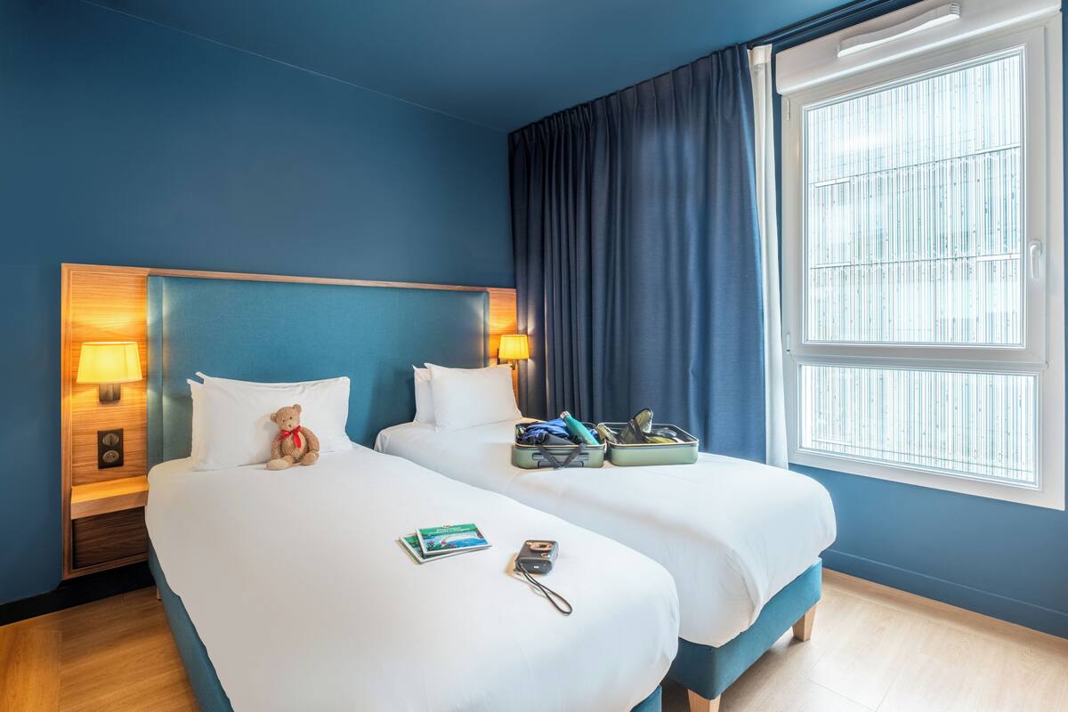 Habitación de hotel con dos camas, una mesita de noche con lámpara, una ventana con persianas y una maleta sobre una cama.