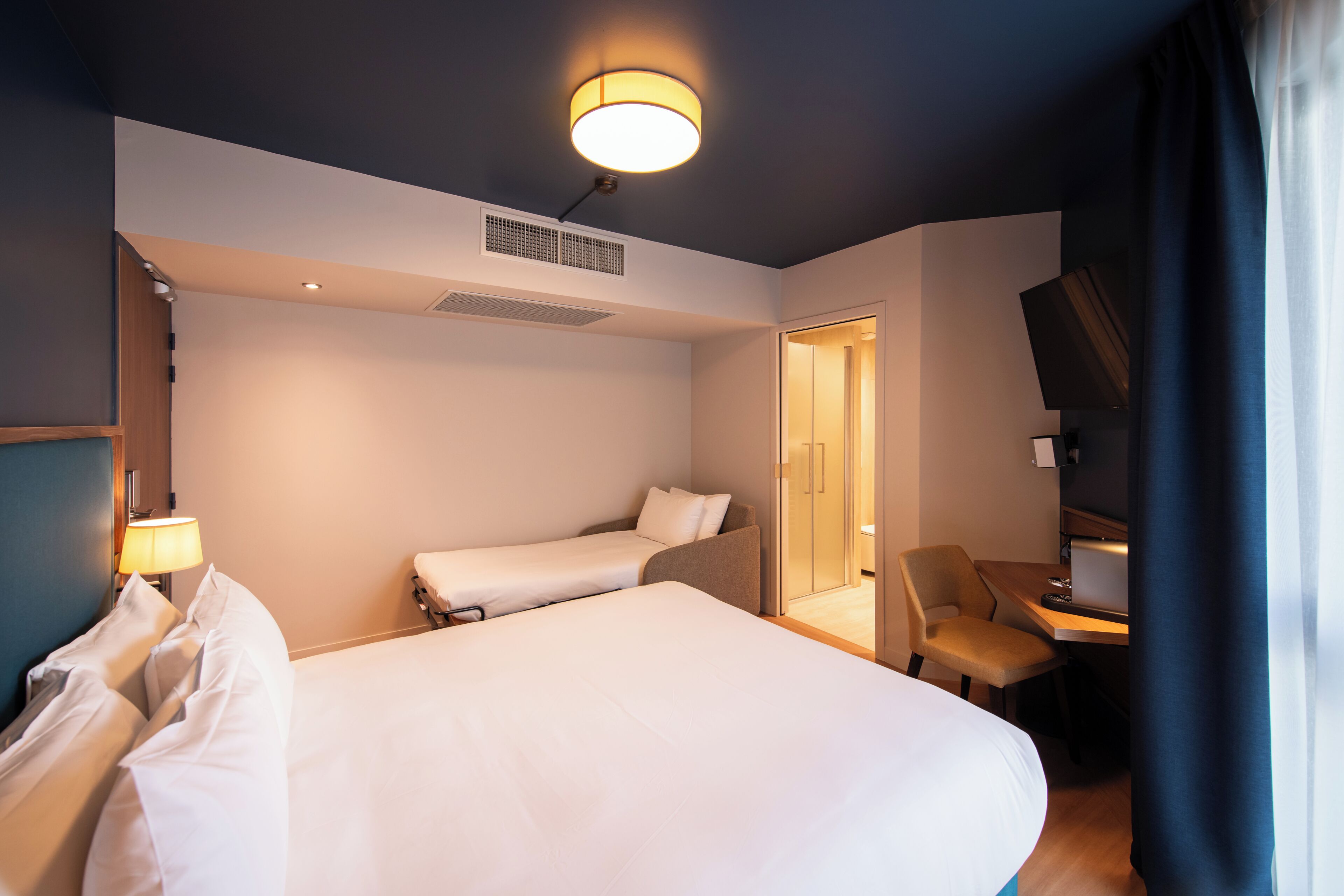 A hotel room with two beds, a desk, a chair, a lamp, and a ceiling light.