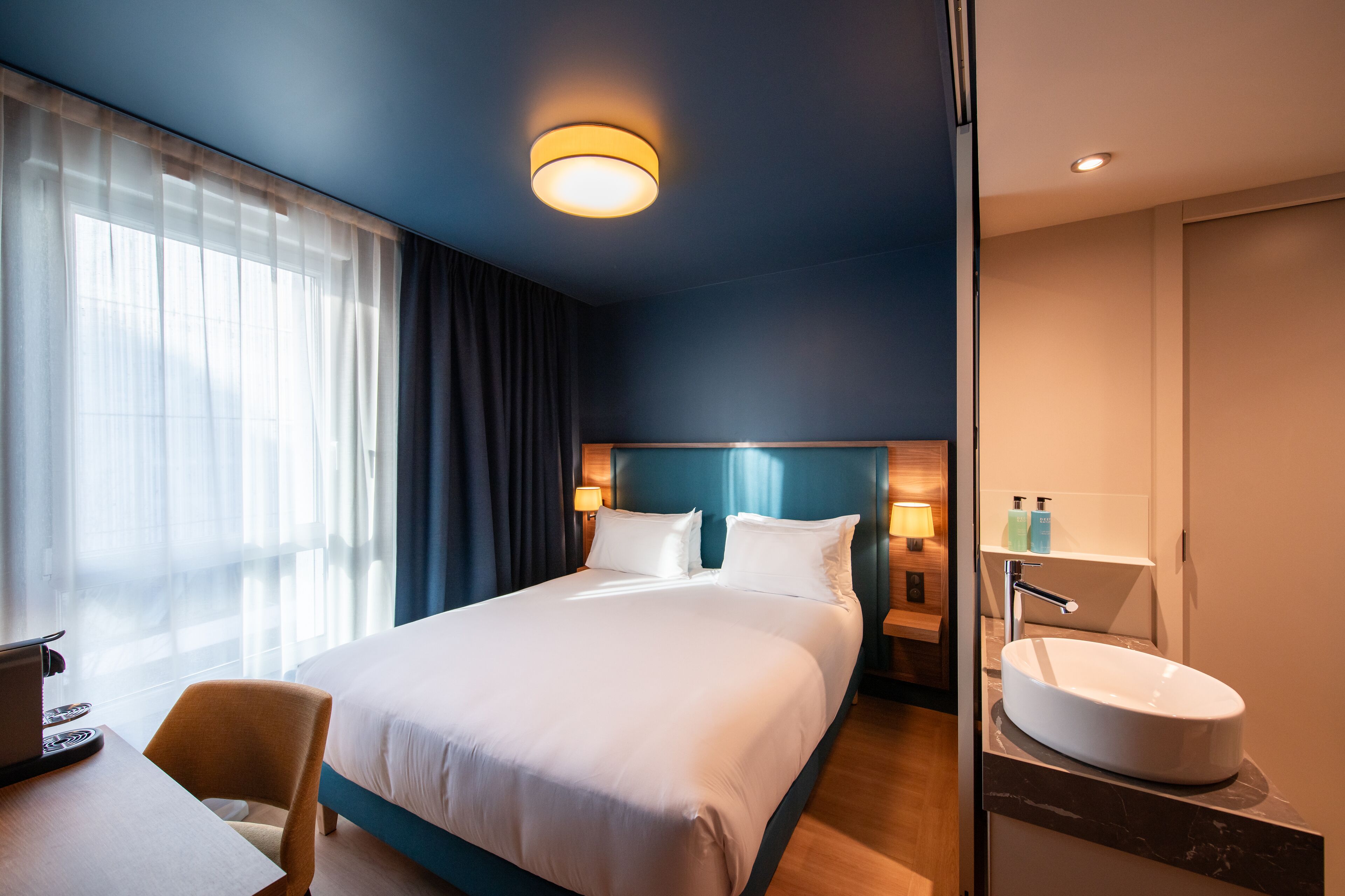 Une chambre d’hôtel avec un grand lit, un bureau avec un ordinateur portable, une salle de bain avec un lavabo et une fenêtre avec des rideaux.