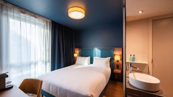 A hotel room with a large bed, a desk with a laptop, a bathroom with a sink, and a window with curtains.