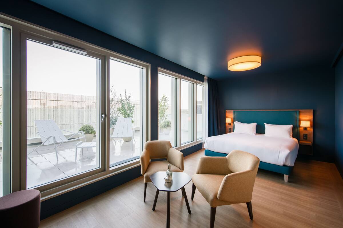 Habitación de hotel con una cama grande, dos sillones, una mesita y vistas al exterior a través de amplios ventanales.