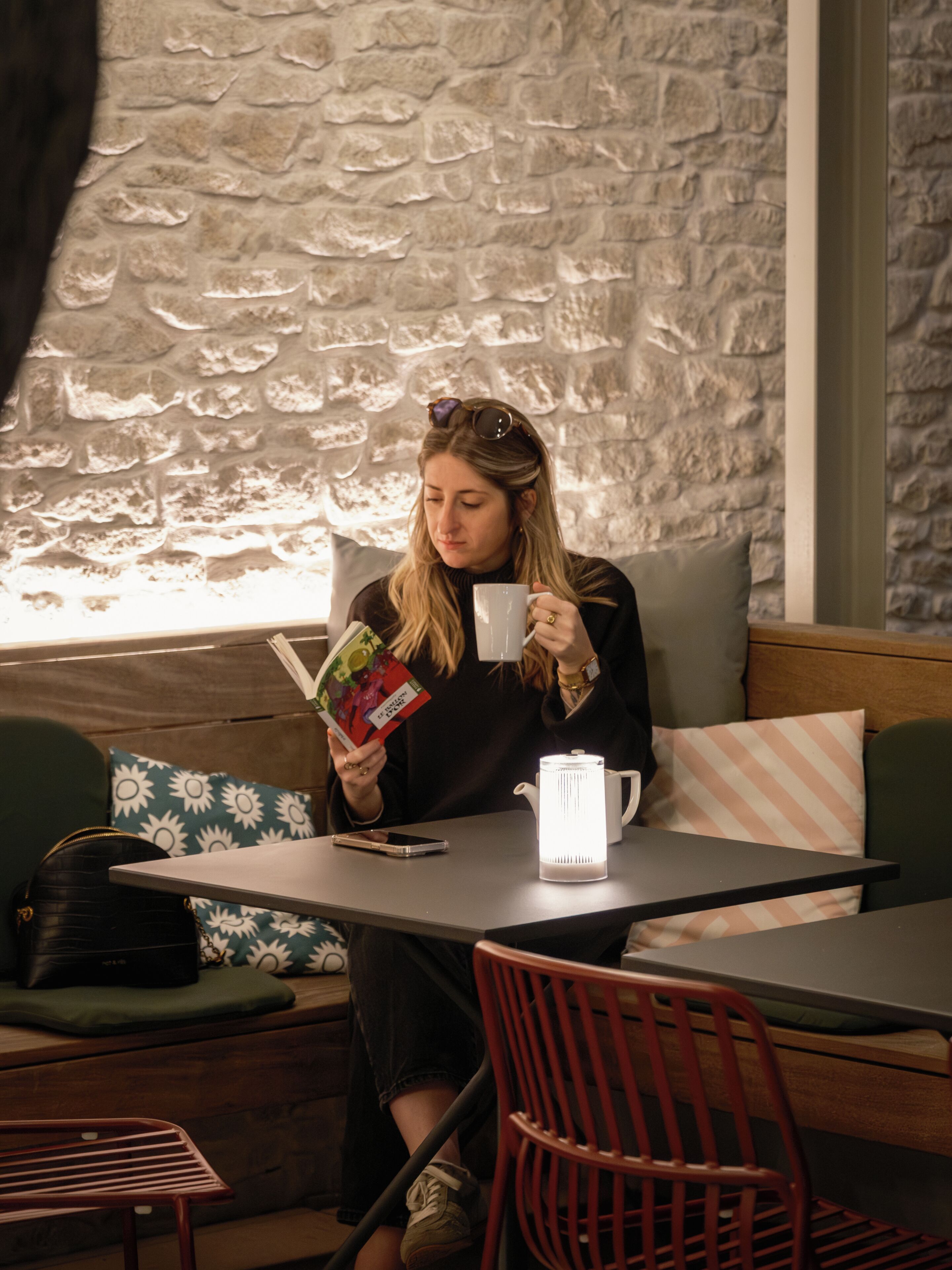 A person seated at a table with a book, a mug, and a kettle. The setting includes a stone wall, a cushioned bench, and a patterned pillow.