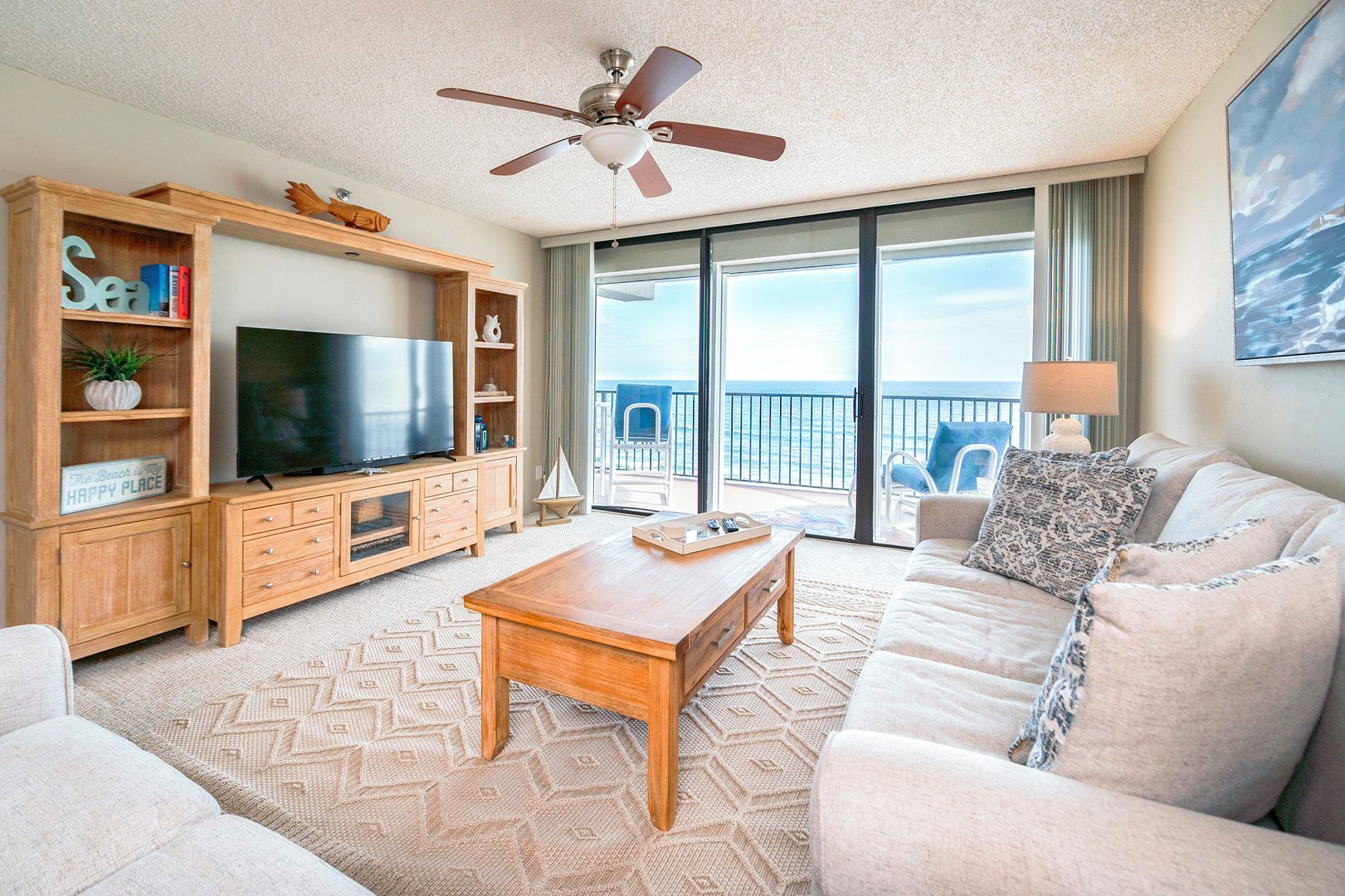 A living room with a television, wooden furniture, a ceiling fan, and a view of the ocean through large windows.
