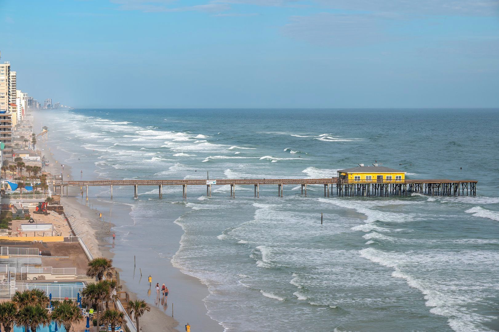 A coastal scene with a pier extending into the ocean, a beach with people, and a yellow building on the pier.