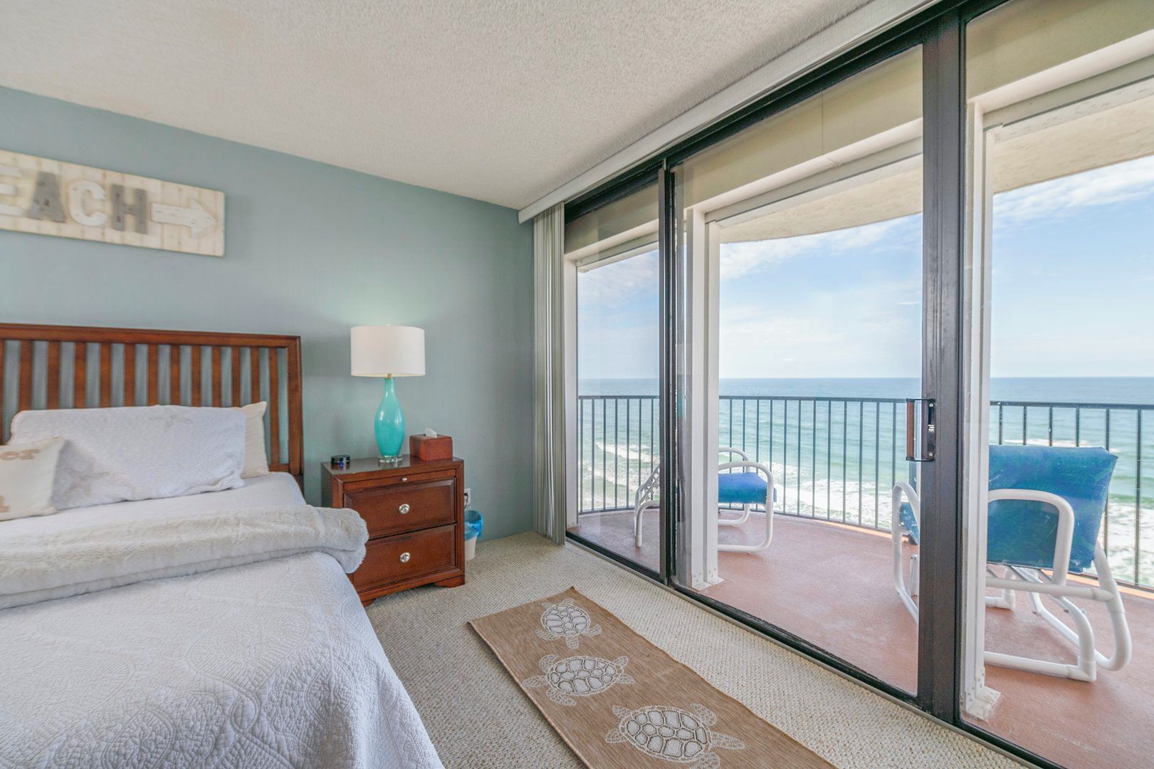 Quarto com cama, mesa de cabeceira, abajur e vista para o oceano através de uma porta de correr de vidro.