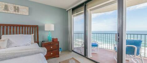 A bedroom with a bed, a nightstand, a lamp, and a view of the ocean through a sliding glass door.
