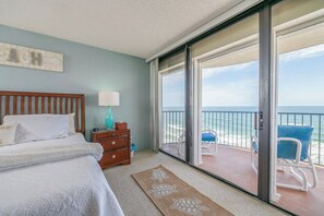 Quarto com cama, mesa de cabeceira, abajur e vista para o oceano através de uma porta de correr de vidro.