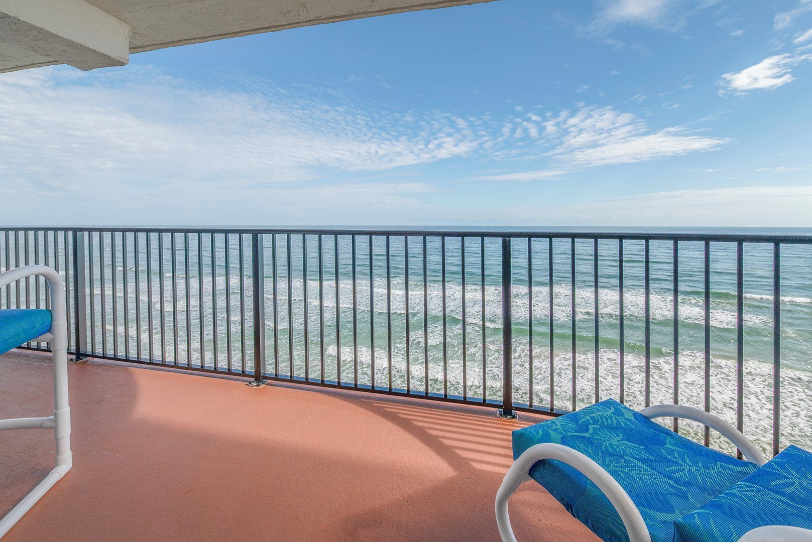 A balcony with a view of the ocean, metal railing, and two chairs with blue cushions.
