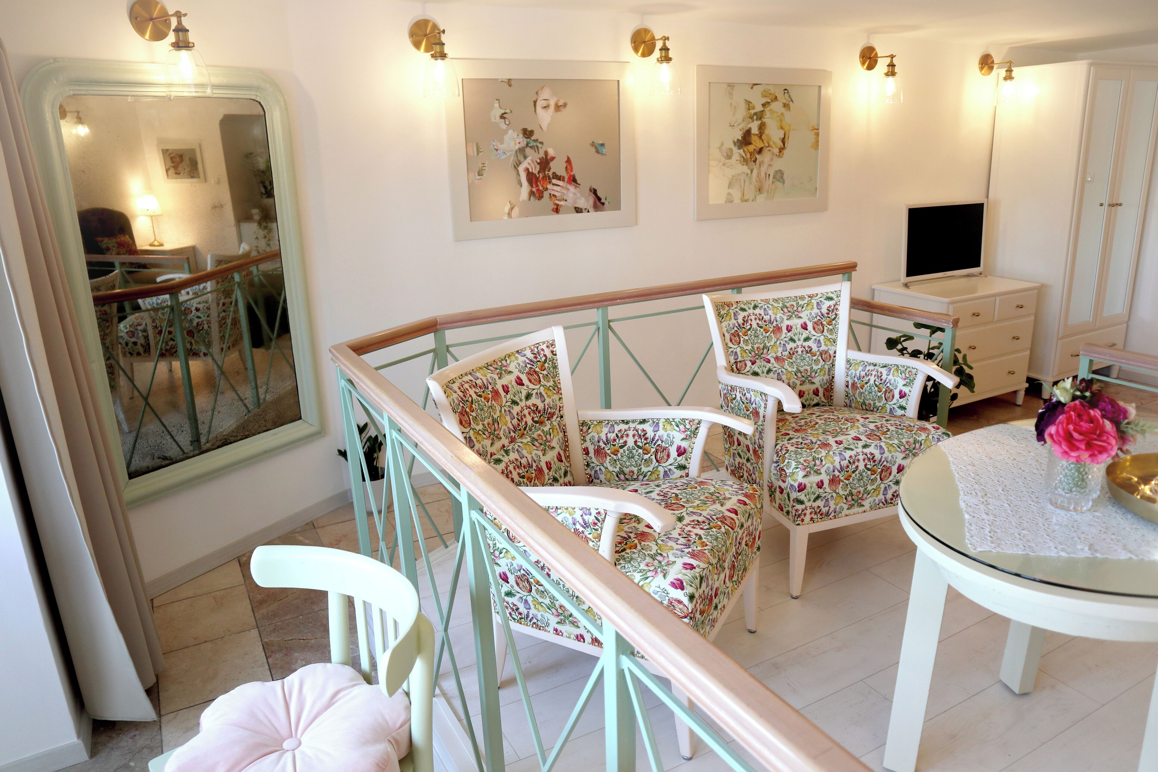 A room with a mirrored wall, floral-patterned chairs, a small round table with a lace tablecloth, and a television on a dresser.