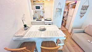 Une cuisine compacte avec un plan de travail blanc, un four à micro-ondes et une cuisinière. On y trouve deux chaises en bois et une petite table sur laquelle est posée une assiette décorative.