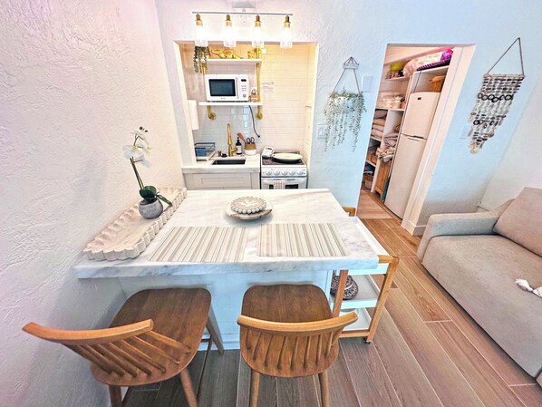 A compact kitchen with a white countertop, a microwave, and a stove. There are two wooden chairs and a small table with a decorative plate.