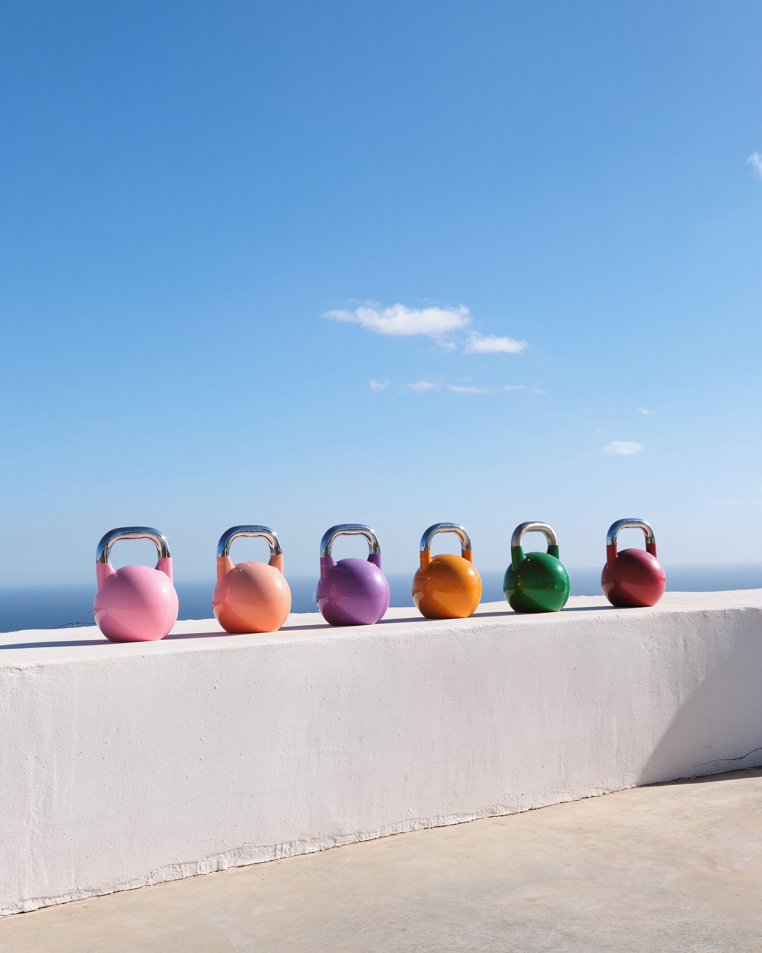 Six kettlebells in various colors lined up on a white ledge with a clear blue sky in the background.