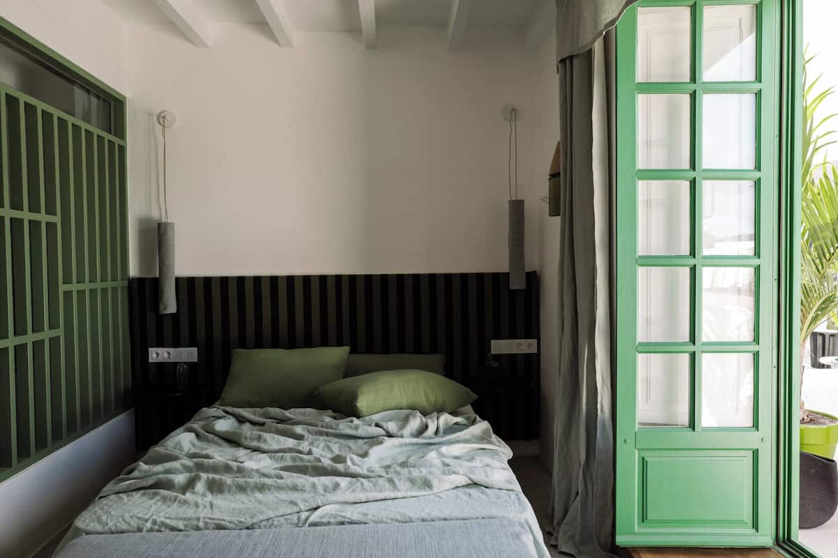 Un dormitorio con cama, mesita de noche, una puerta verde y ventana con cortinas.