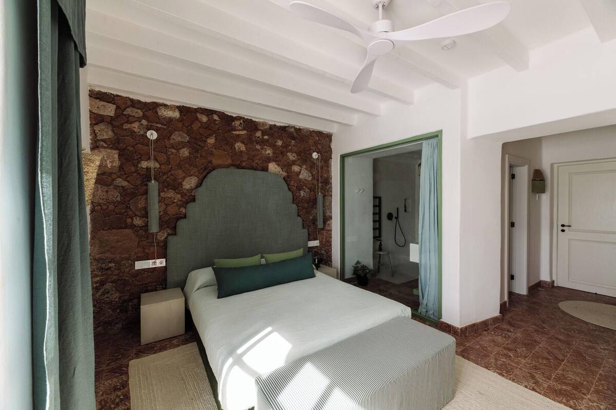Un dormitorio con pared de piedra, cama con almohadas verdes, ventilador de techo y baño visible a través de una puerta abierta.