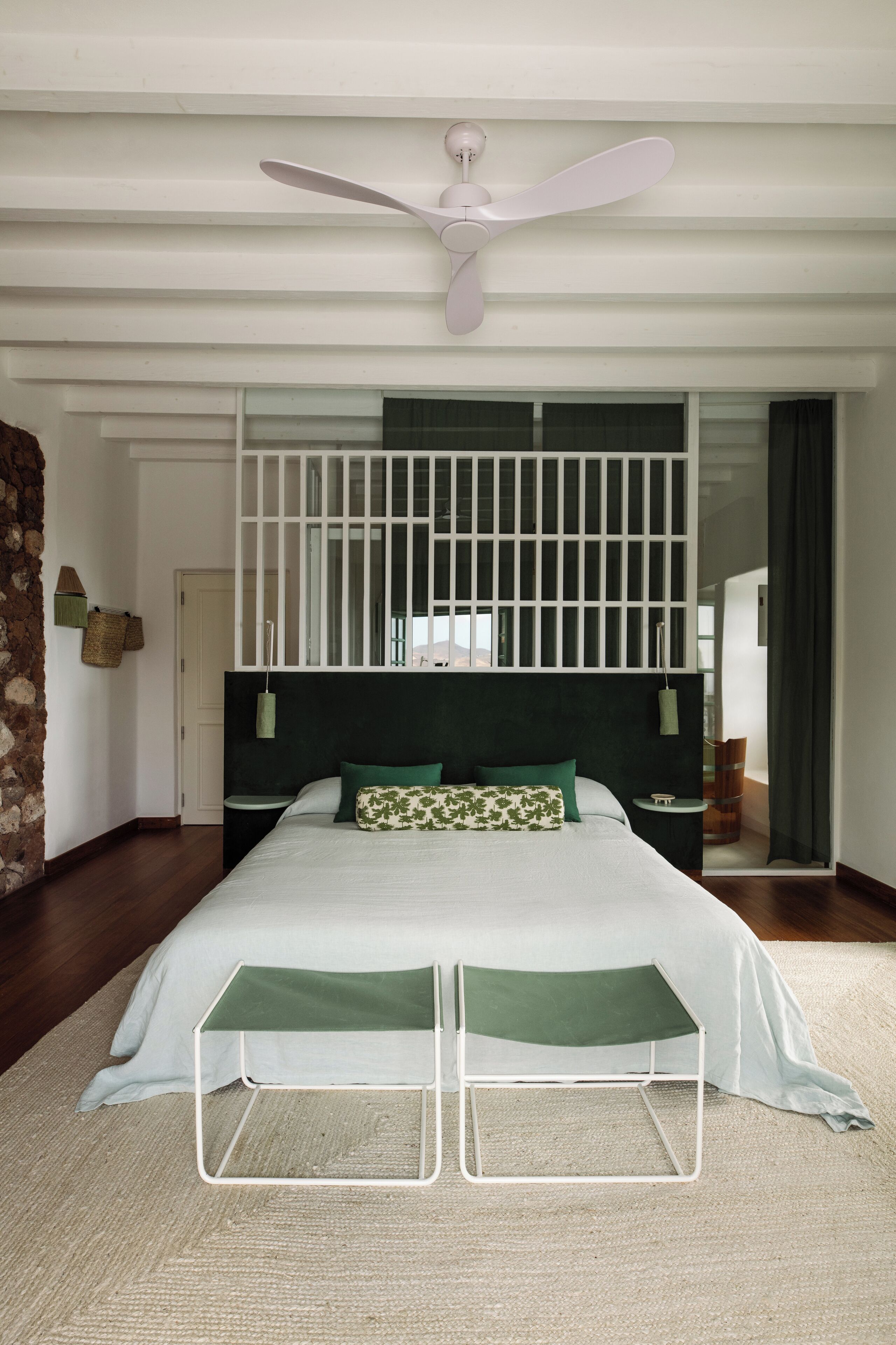 A bedroom with a bed, a standing fan, a nightstand, and a window with a view.