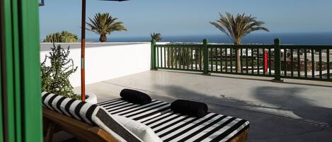 A balcony with a lounge chair and an umbrella, overlooking a coastal view with palm trees and a fence.