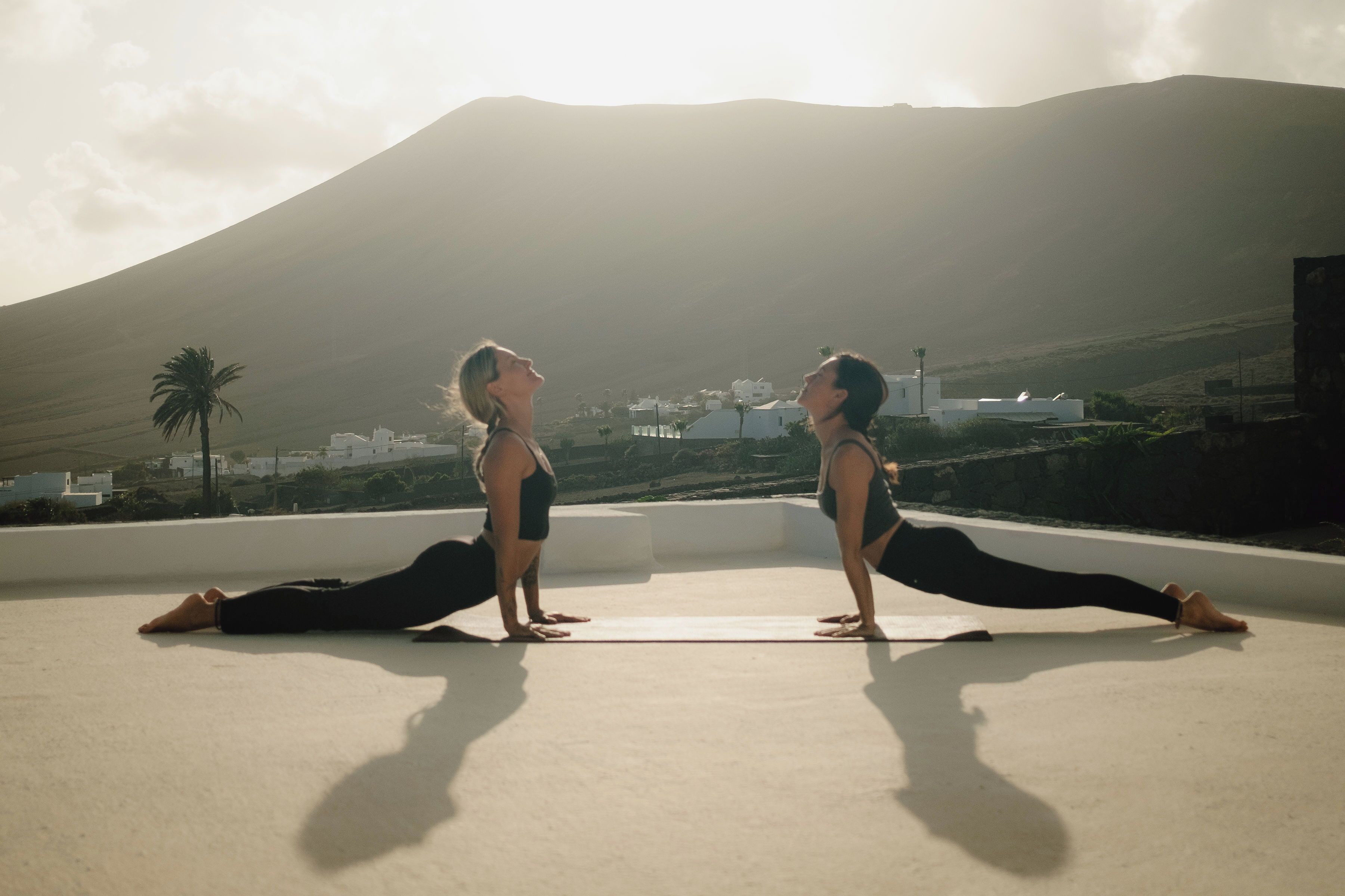 Dos personas realizan una postura de yoga en una azotea, con una montaña al fondo.