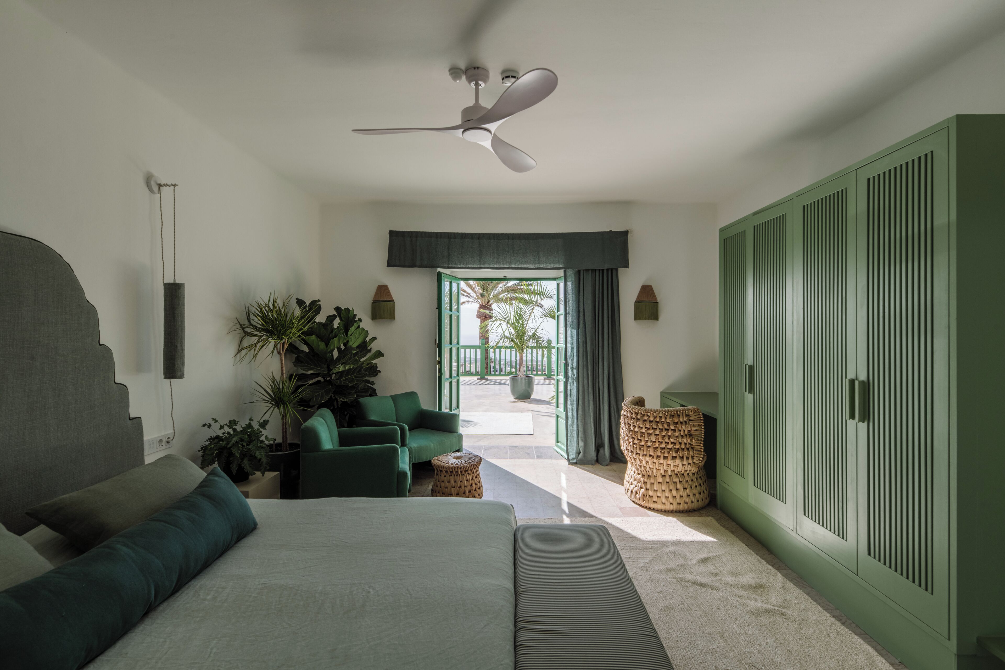 A bedroom with a bed, green armchair, potted plants, a ceiling fan, and a view to the outside.