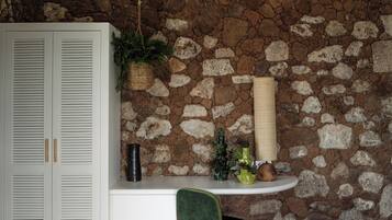 A room with a stone wall, white ceiling beams, a bed, a green chair, a small table with plants, and a white wardrobe.