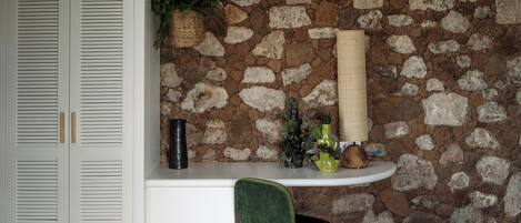 Una habitación con pared de piedra, vigas de techo blancas, una cama, una silla verde, una mesita con plantas y un armario blanco.