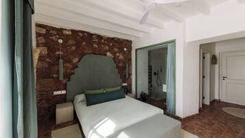 Un dormitorio con pared de piedra, una cama con almohadas verdes, un ventilador de techo y una puerta de baño visible.