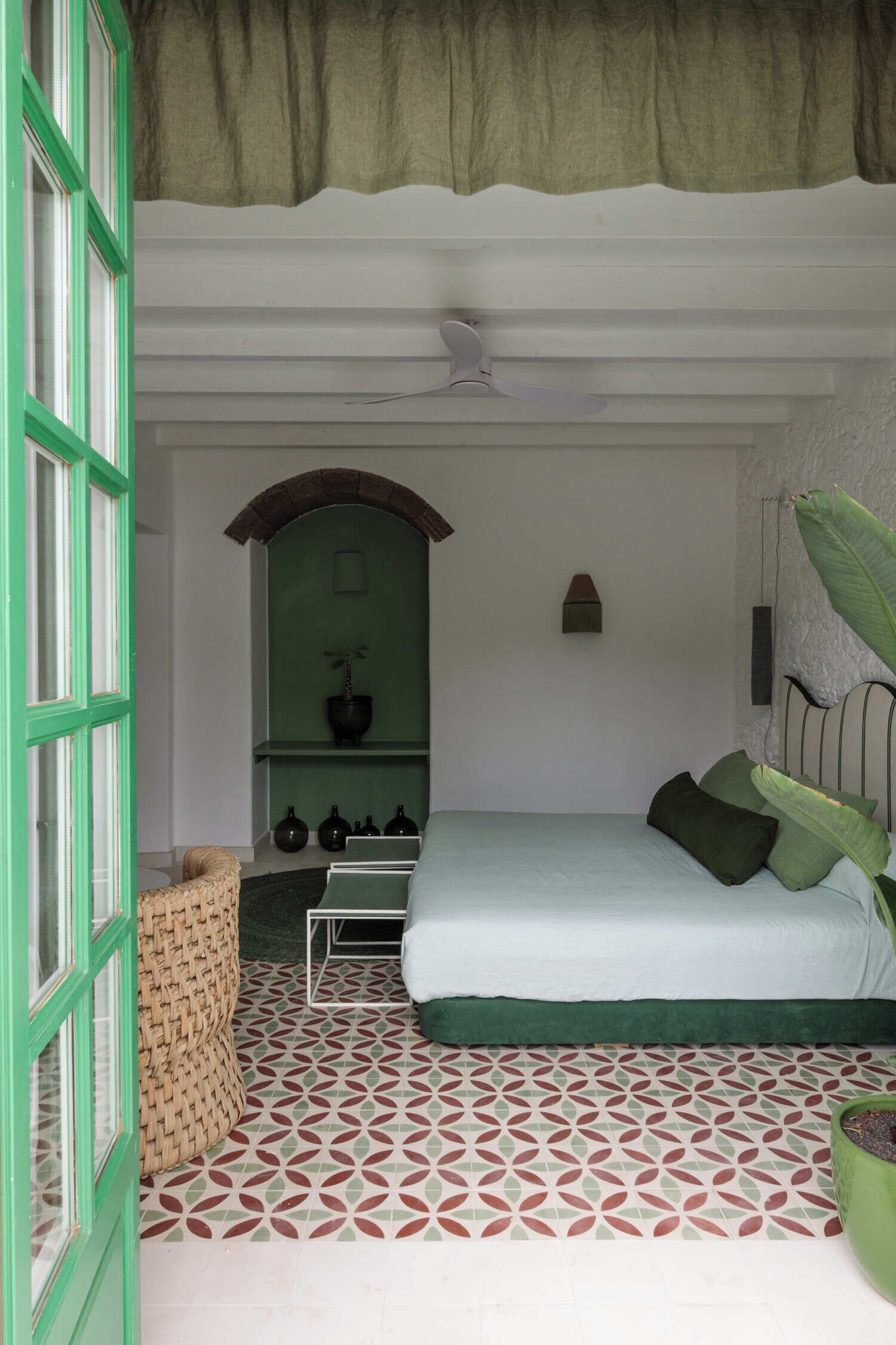 Un dormitorio con una cama, almohadas verdes, una planta en maceta, un estante con adornos y un suelo estampado.