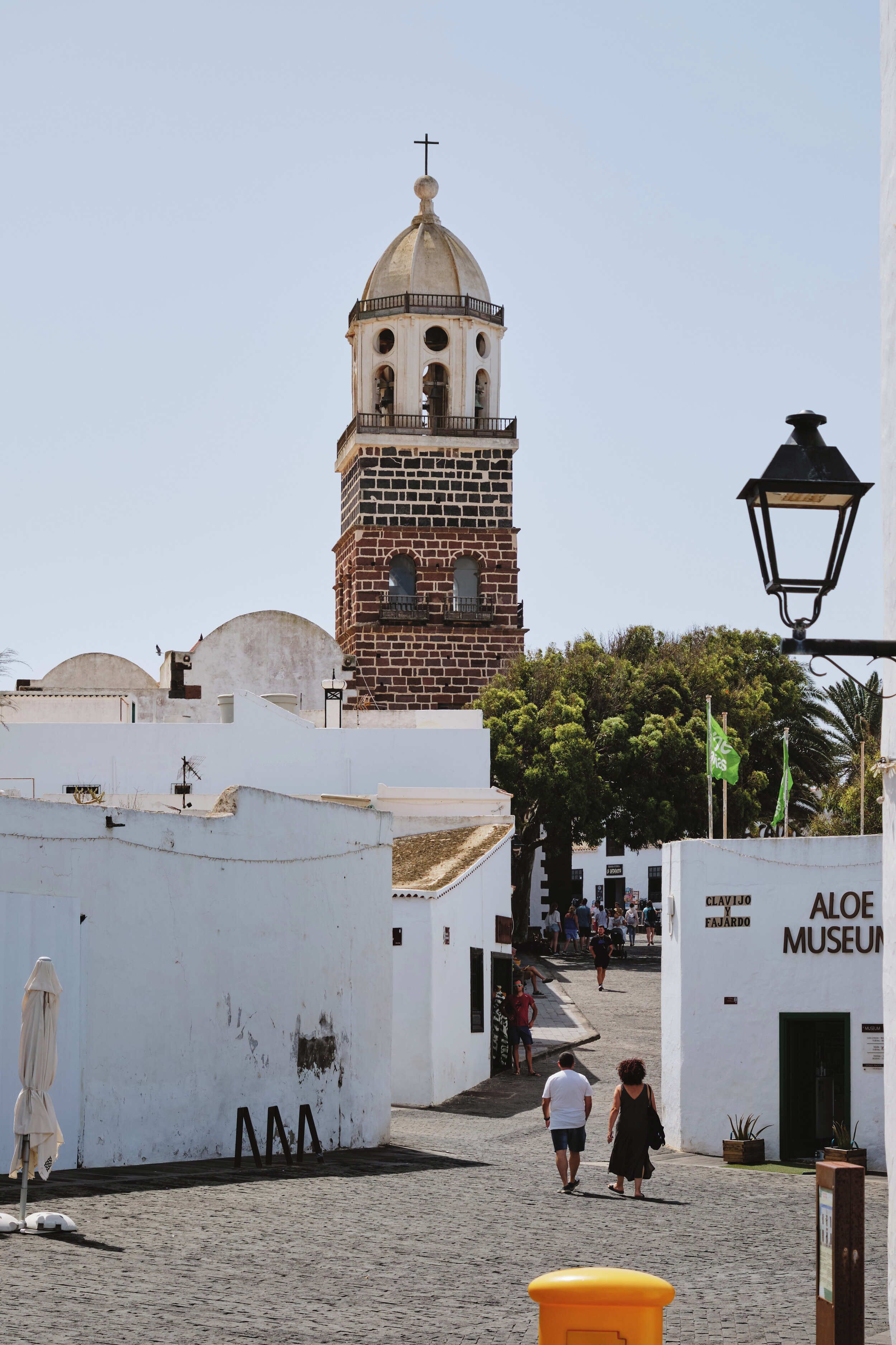 Una calle adoquinada con una torre histórica, un edificio blanco que dice "Aloe Museum" y un farol.