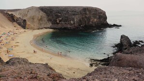 A beach with a large rock formation, clear turquoise water, and people sunbathing.