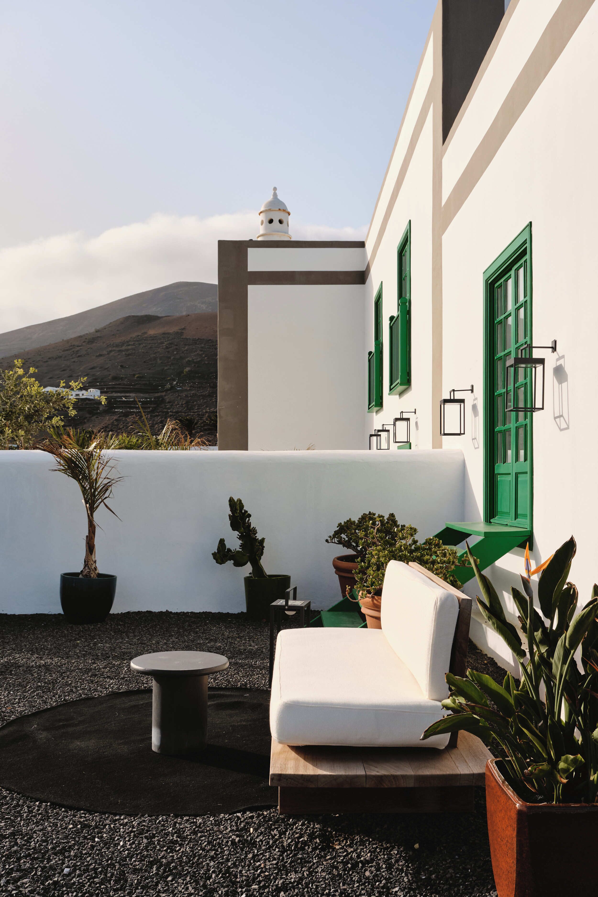 A white building with green shutters and a white wall, a white sofa, a small round table, and potted plants.