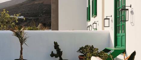 A white building with green shutters and a white wall, a white sofa, a small round table, and potted plants.
