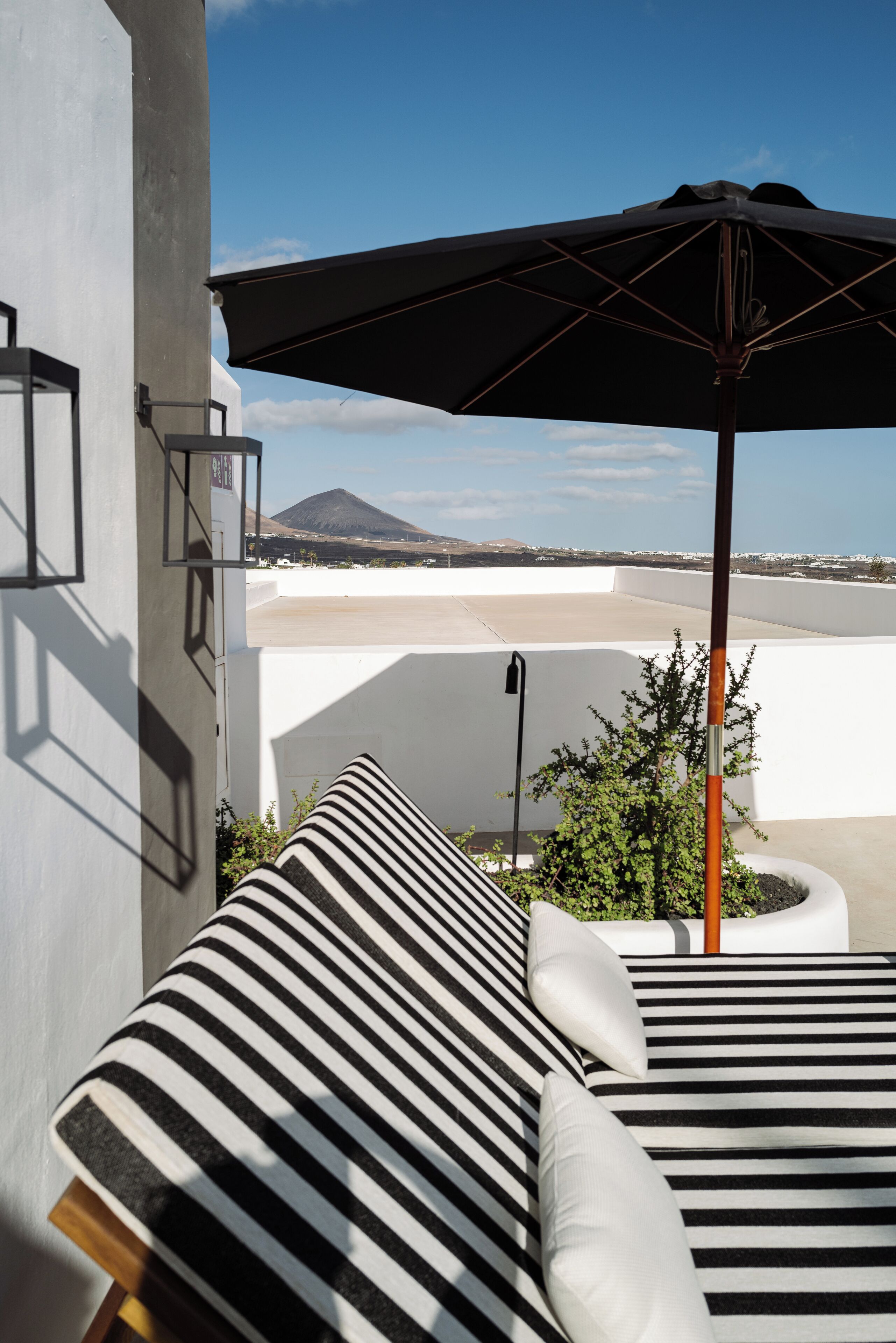 A modern outdoor lounge area with striped cushions and a large umbrella, overlooking a beach and a mountain.