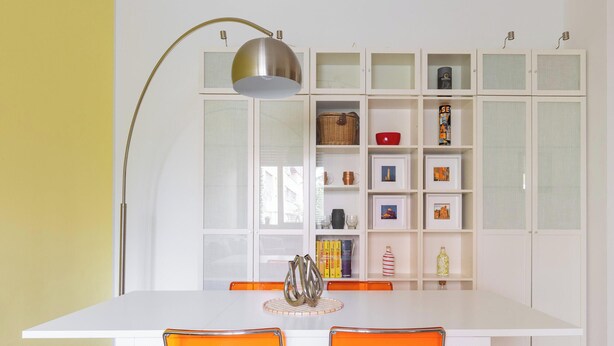 Een eetruimte met een witte tafel, oranje stoelen en een witte kast met glazen deuren op de achtergrond.