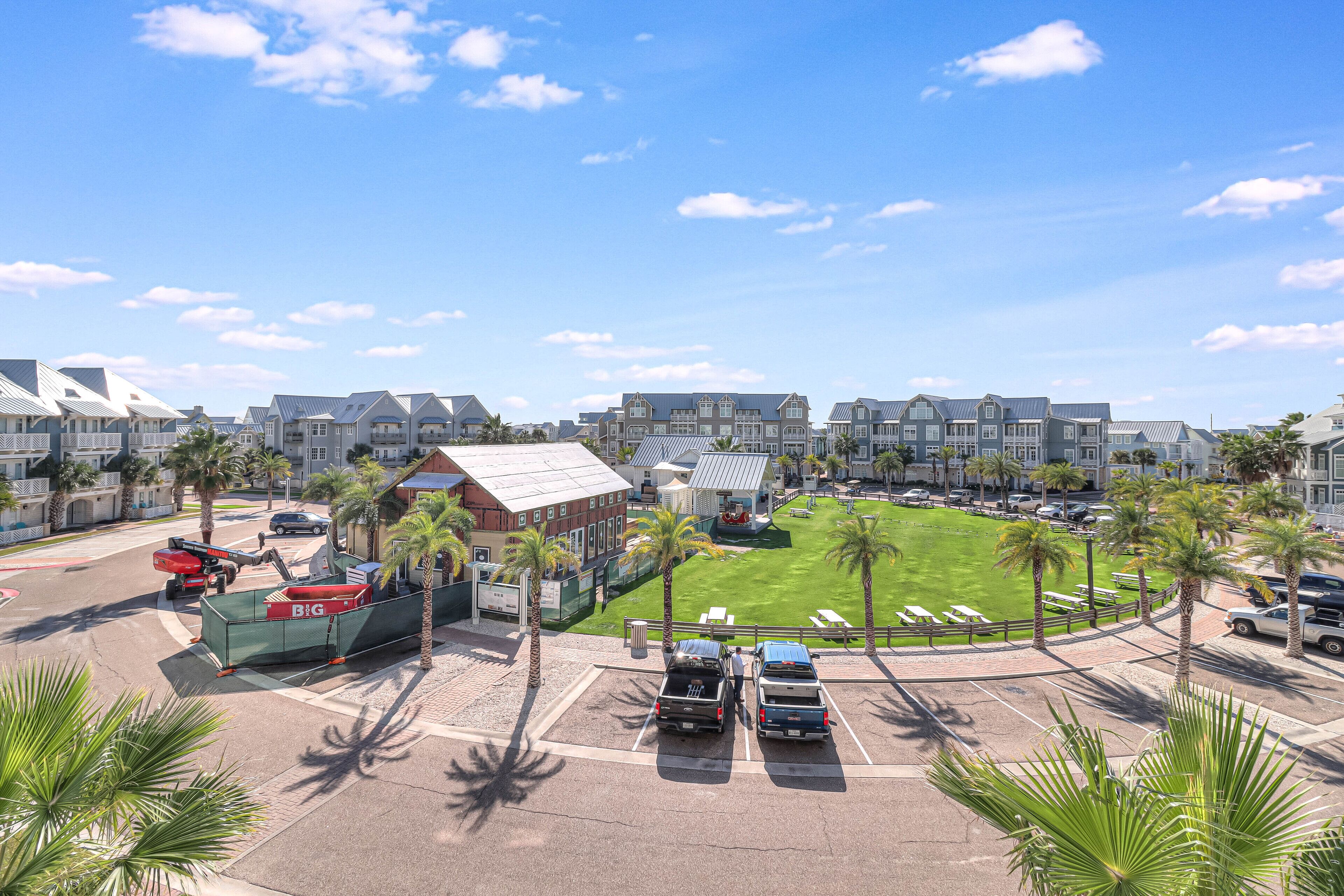 A parking lot with several vehicles, a grassy area with palm trees, and buildings in the background.