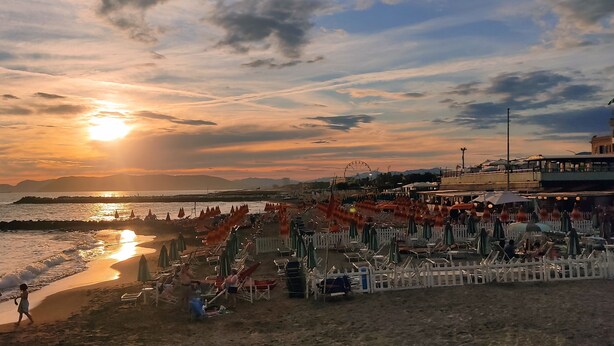 Ein Strand mit Sonnenschirmen und Liegestühlen, einem Steg und einem Riesenrad im Hintergrund.