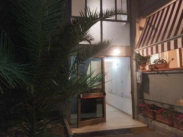 A narrow outdoor corridor with a glass door leading to a room, surrounded by potted plants and a striped awning.