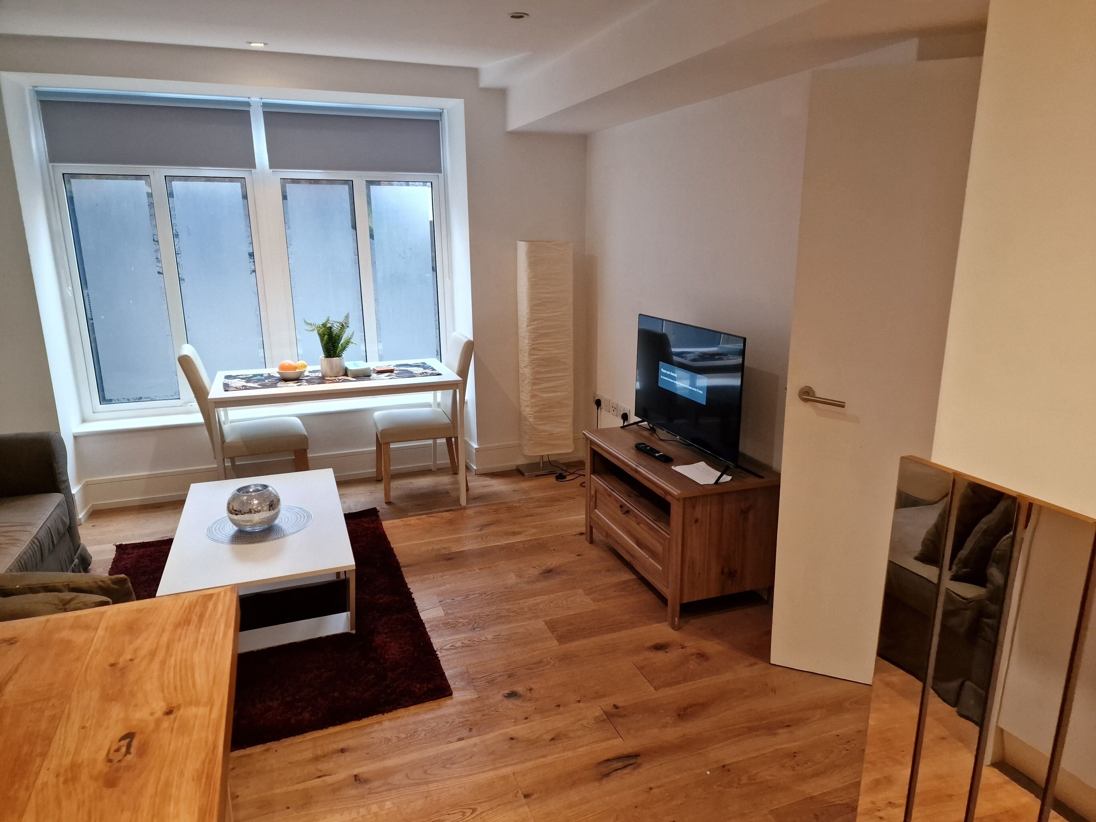 Een moderne woonkamer met een houten vloer, een witte bank, een eettafel met stoelen, een tv op een houten dressoir en een raam met geëtste glas.