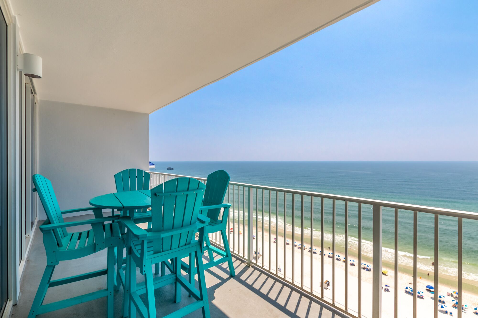 Un balcon avec des chaises turquoise et une table, donnant sur une plage parsemée de parasols et animée par la présence de nombreuses personnes.