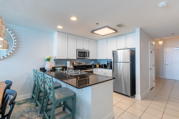 A modern kitchen with white cabinets, stainless steel appliances, and a central island with bar stools. - Lighthouse 1507 by Liquid Life (Gulf Shores)