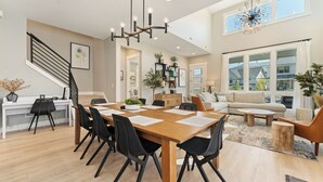 A spacious living area with a wooden dining table, black chairs, a sofa, and a coffee table. There is a staircase with a black railing and a large window with a view of the outside. - Bighorn & Bluebell at Chelan Lookout Vacation Rentals (Chelan)