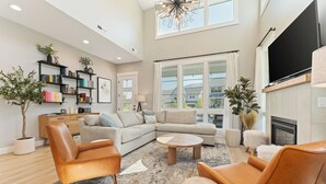 A spacious living room with a large sectional sofa, two leather armchairs, a coffee table, and a fireplace. There is a bookshelf with books and decorative items, and a large window offering a view of the outside. - Bighorn & Bluebell at Chelan Lookout Vacation Rentals (Chelan)