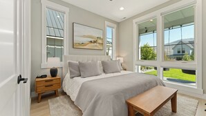 A bedroom with a large bed, a nightstand, a bedside table, a bench, and a window with a view of a house and greenery. - Bighorn & Bluebell at Chelan Lookout Vacation Rentals (Chelan)