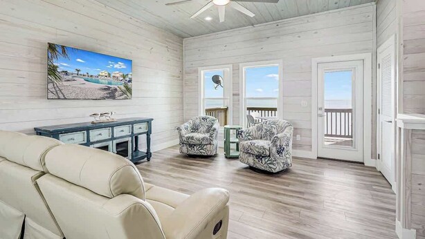 Una habitación con ventilador de techo, un cuadro de playa, dos sillones estampados, un sofá y vistas por una puerta.