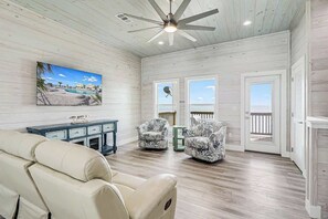 Una habitación con ventilador de techo, un cuadro de playa, dos sillones estampados, un sofá y vistas por una puerta.