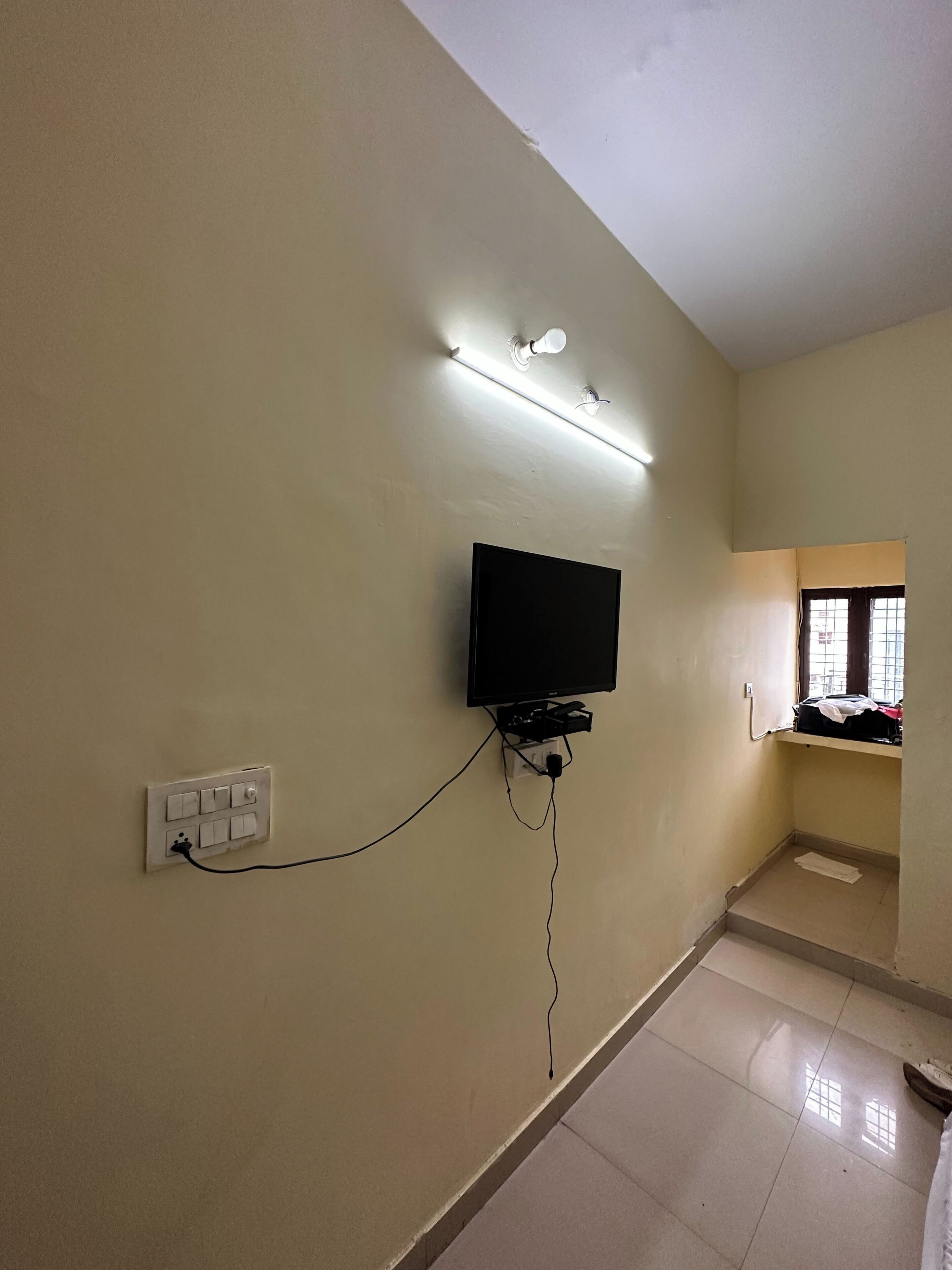 Ein Zimmer mit einem an der Wand befestigten Fernseher, einer Steckdose und einem Fenster mit Vorhängen.