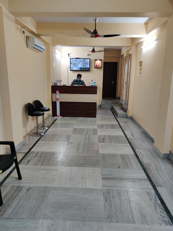 A reception area with a reception desk, chairs, a ceiling fan, a TV, and a tiled floor. - Sanjiva Suites (Barasat)