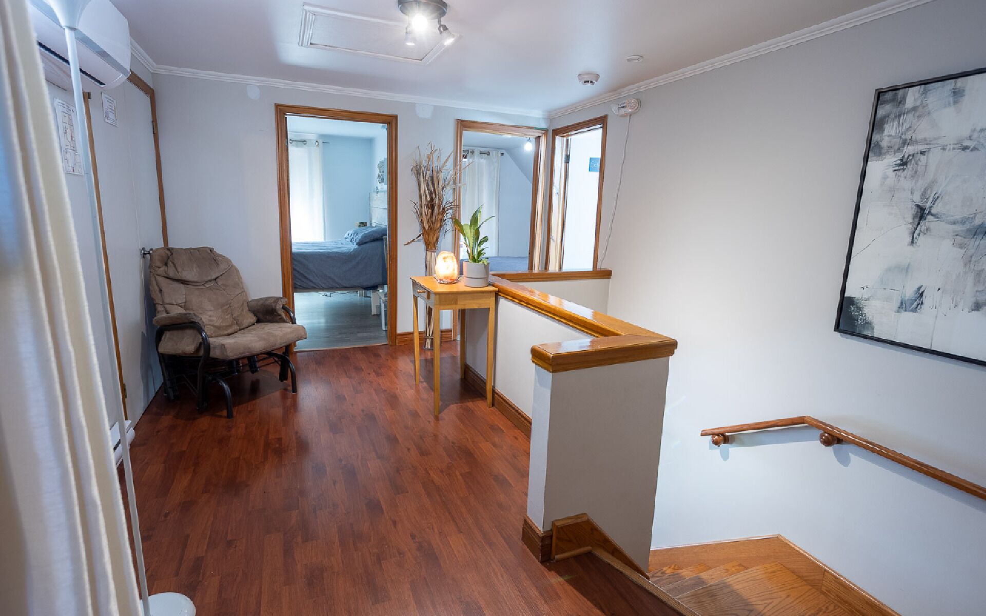 A spacious room with hardwood flooring, a wooden chair, a small table with a vase and plant, a staircase with a wooden handrail, and a large abstract painting on the wall.