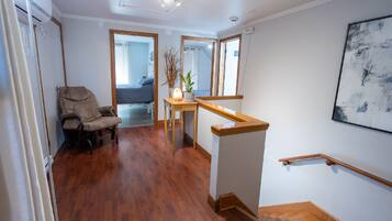 A spacious room with hardwood flooring, a wooden chair, a small table with a vase and plant, a staircase with a wooden handrail, and a large abstract painting on the wall.