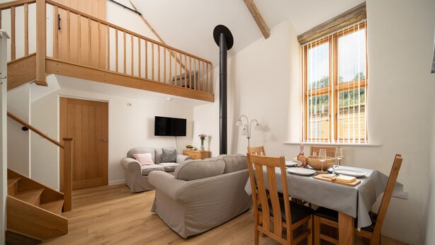 A spacious living area with a wooden staircase, a sofa set, a dining table with chairs, and a large window with a view.