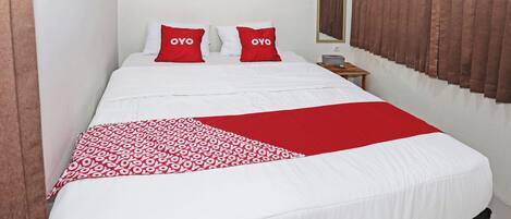 A neatly made bed with white and red bedding, a bedside table with a lamp, and a window with brown curtains.